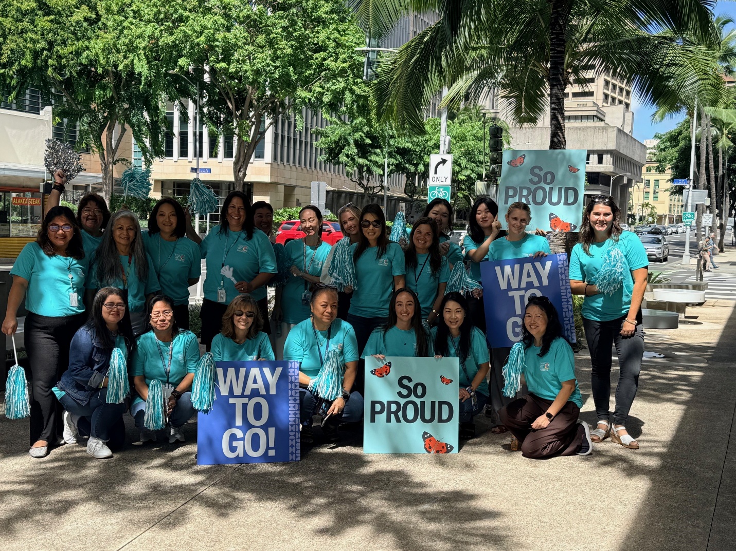 Women in teal shirts from Central Pacific Bank Foundation hold signs and pom poms in solidarity with Men's March participants.