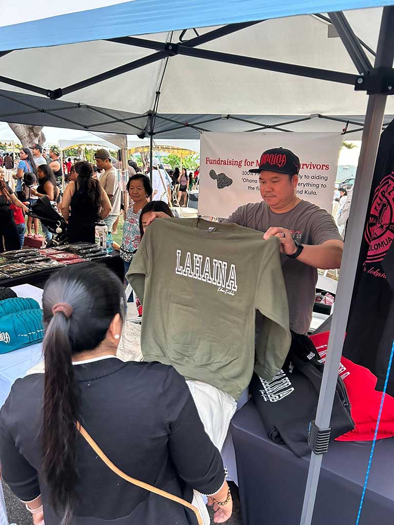A male vendor holds up an olive sweatshirt for sale with to raise funds for Maui wildfire survivors.